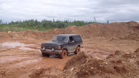1987 Ford Bronco - A dark-colored Ford Bronco is navigating through a muddy, off-road environment with large piles of dirt and trees in the background.