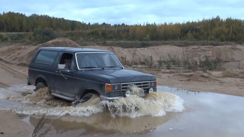 1987 Ford Bronco - A dark-colored Ford Bronco is driving through a shallow body of water, creating large splashes. The vehicle is off-road in a sandy area wit…