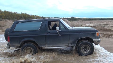 1987 Ford Bronco - A dark-colored Ford Bronco is driving through murky water in a sandy, outdoor environment. The vehicle is viewed from the side, showing its…