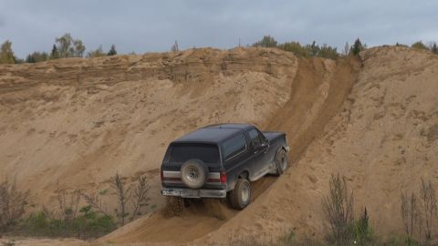 1987 Ford Bronco - A black Ford Bronco is driving up a steep sand dune, kicking up dirt from its tires. The vehicle is seen from a rear three-quarter angle ag…