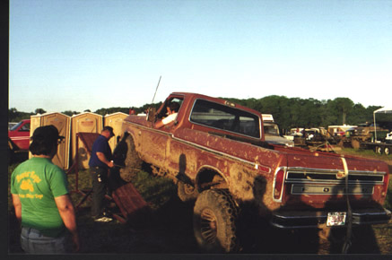 1991 Ford Bronco - A muddy, lifted red Ford pickup truck is the focus, with portable toilets and other vehicles in the background.