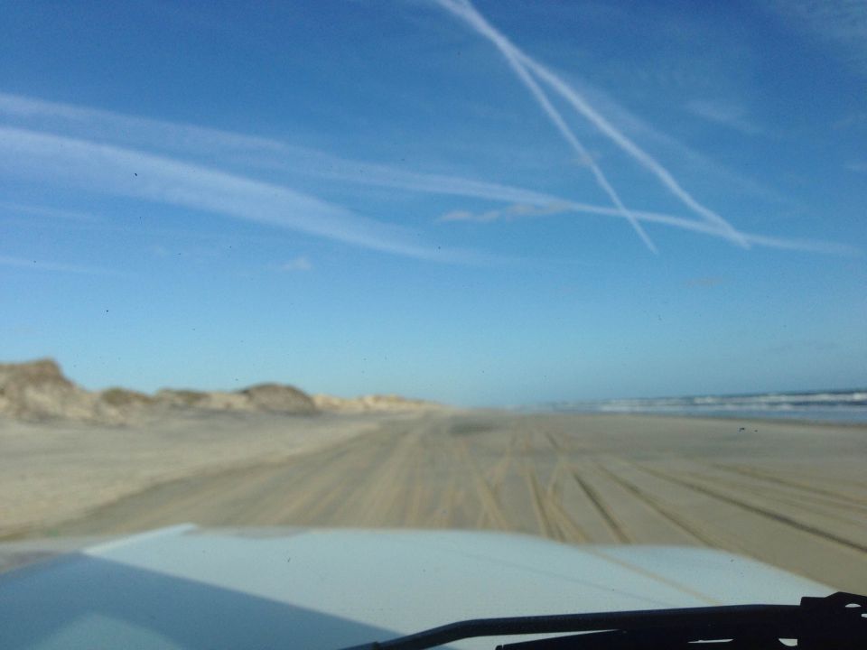 View from inside a vehicle driving on a beach. Dunes on the left, ocean on the right, blue sky with contrails.