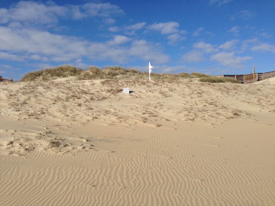 Sandy dunes with a white flag and wooden walkway under a blue sky.
