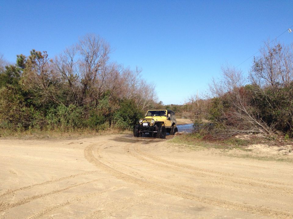 Yellow Jeep Wrangler driving through water on a sandy path.