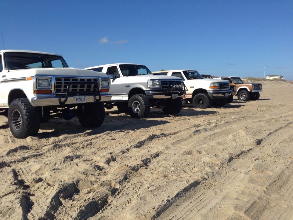 Four lifted Ford trucks and SUVs parked on a sandy beach.