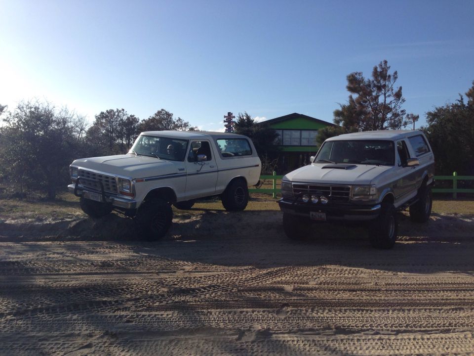 Two Ford Broncos parked on sand, one white and older, one silver and newer.