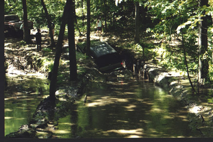 1991 Ford Bronco - Two vehicles are present in a wooded area, one stuck in a water-filled ditch.