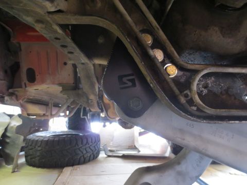 1996 Ford Bronco - Underneath view of a vehicle's frame, showing suspension components, a spare tire, and a metal skid plate with a "S" logo. Visible text on…