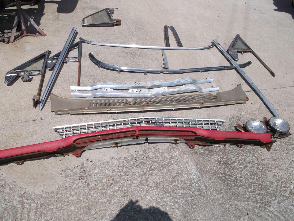 Vintage car parts including red bumper, chrome grille, headlights, and trim pieces on concrete.