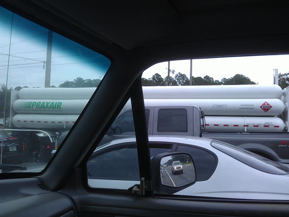 View from inside a vehicle at a truck with Praxair hydrogen tanks. Foreground shows a white car.