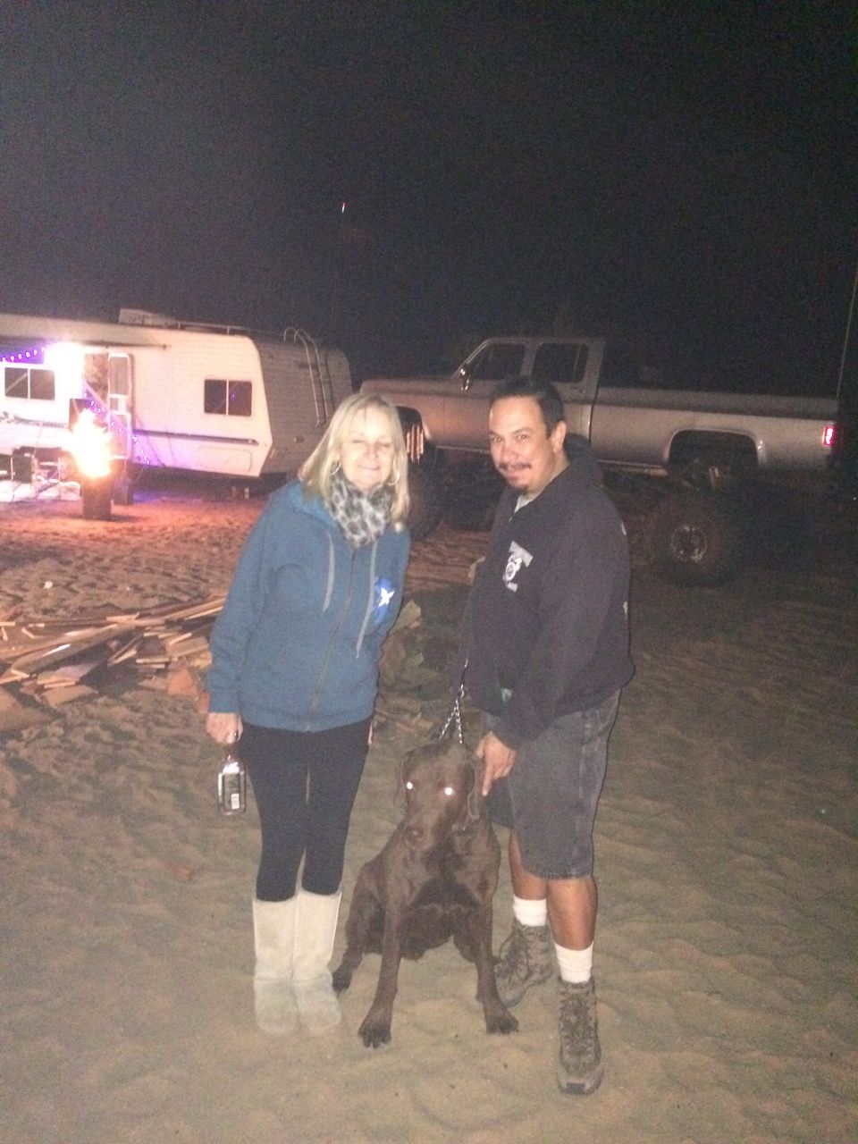 A man, woman, and dog pose in front of a trailer and pickup truck at night.