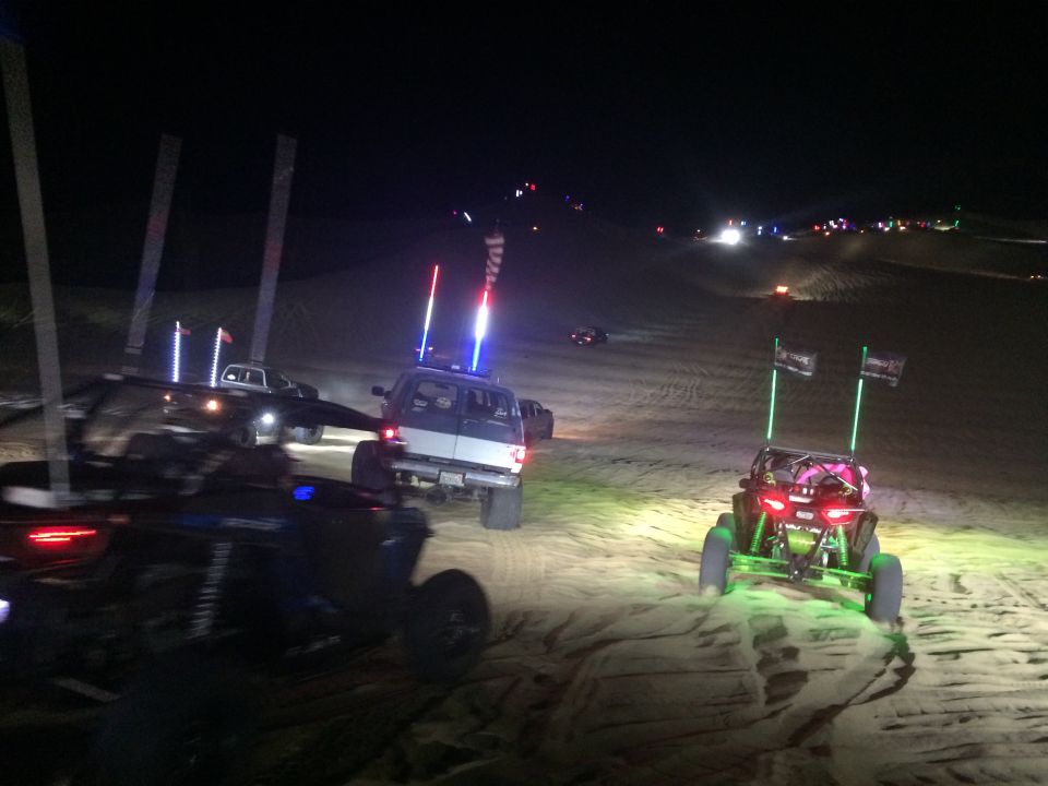 Off-road vehicles with lights driving on sand dunes at night.