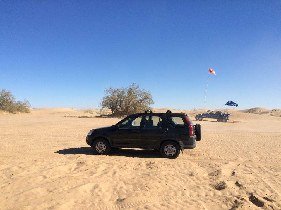 Black Honda CR-V parked in desert dunes with a blue dune buggy in the distance.