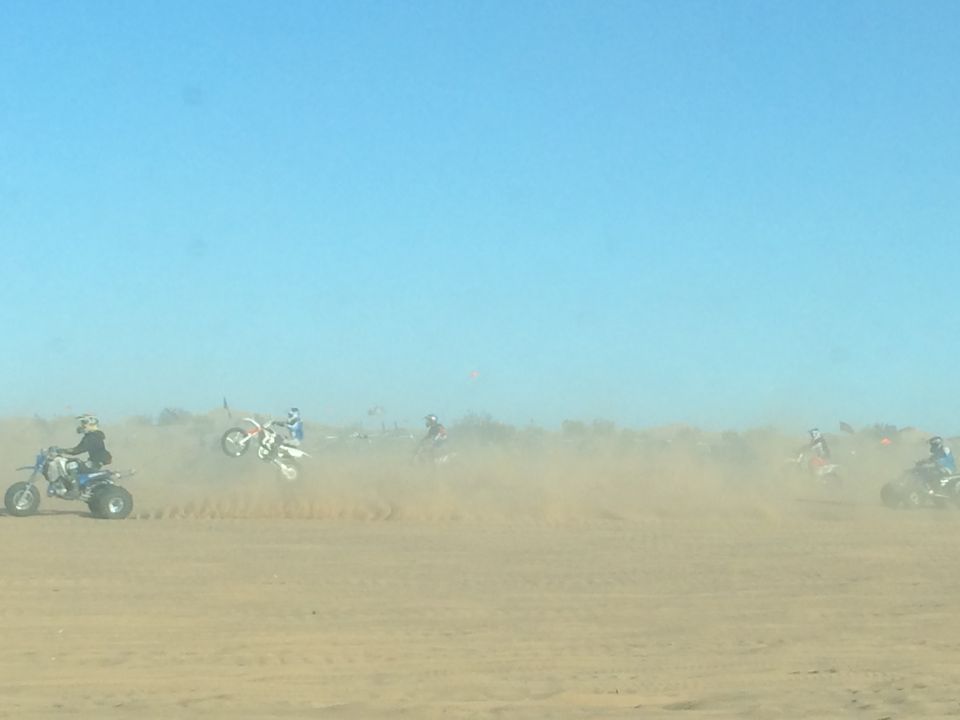 Dirt bikes and ATVs riding on sand dunes, kicking up dust.