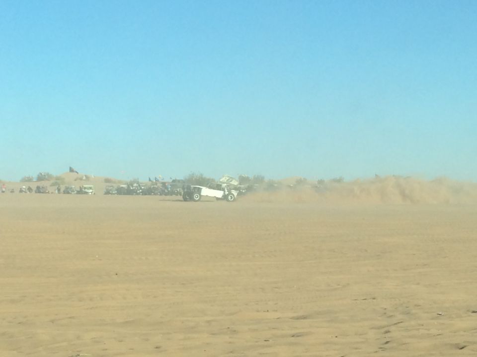 White off-road vehicle driving and kicking up dust in a desert with other vehicles in the background.