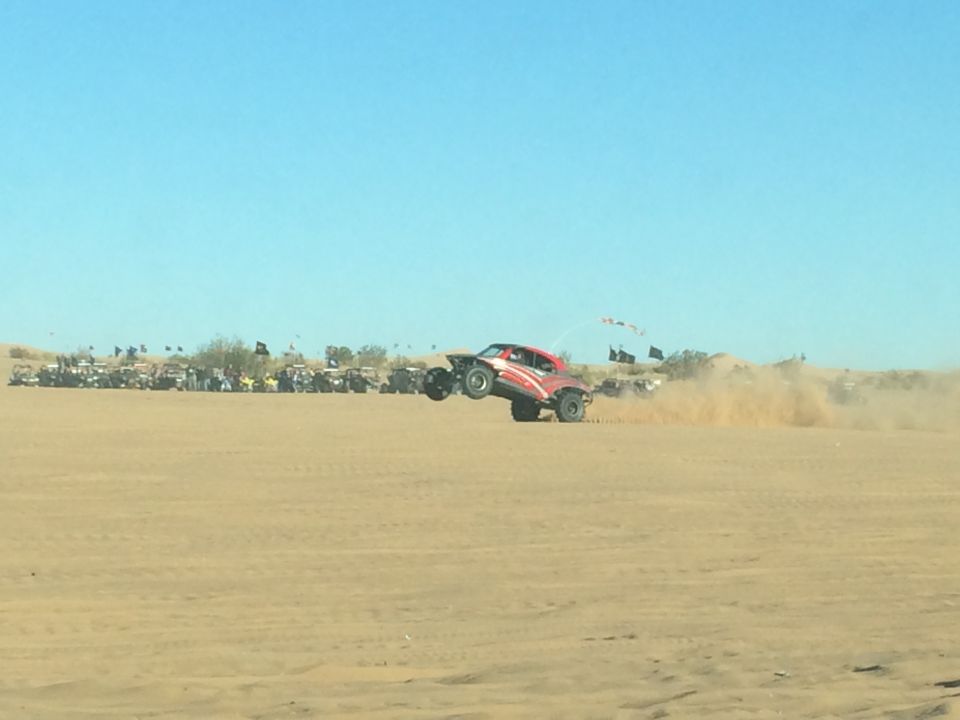 Red and white off-road buggy jumping in the desert, kicking up dust. Parked vehicles in background.