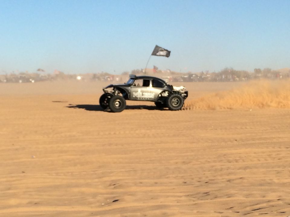 Black off-road buggy with flag kicking up dust on sand dunes.