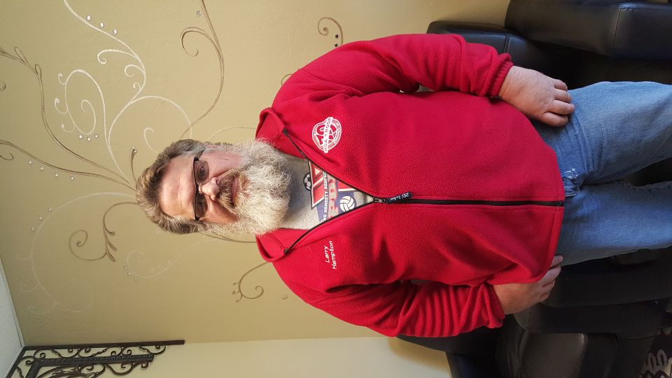 Man with beard and glasses wearing a red jacket and blue jeans.