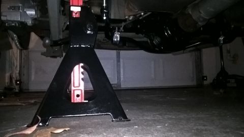 2004 Ford Crown Victoria - The underside of a vehicle is supported by a black jack stand in a garage. The rear differential and exhaust system are visible.