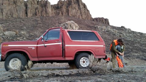 1981 Ford Bronco - A red 1980s Ford Bronco is parked on a dirt road with a barren, rocky hillside in the background. Two people wearing orange vests are stand…