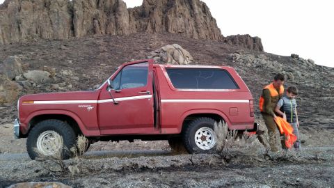 1981 Ford Bronco - A red 1980s Ford Bronco is parked on a dirt road with a man and a boy standing next to it. The background shows a rocky, barren hillside wi…