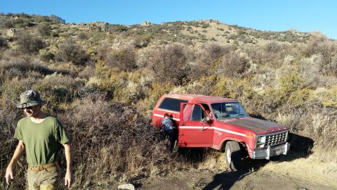 1981 Ford Bronco - A red Ford Bronco is parked on a dirt path in a dry, brushy landscape with a person entering the driver's side. A man in a bucket hat and g…