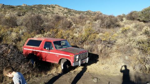 1981 Ford Bronco - A red 1980s Ford Bronco is parked on a dirt path surrounded by dry brush and hills under a clear blue sky. A young boy stands to the left o…
