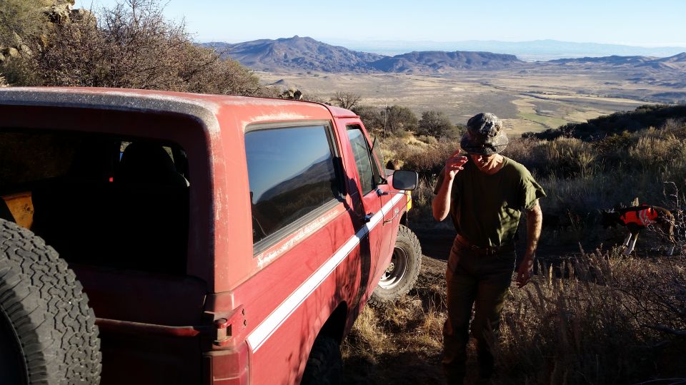 Red 1980s Ford Bronco parked on a dirt road with a man and dog, overlooking a valley.