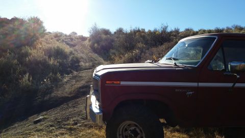 1981 Ford Bronco - A red 1980s Ford Bronco is parked on a dirt road with a steep hill to the left. The sun is shining brightly in the upper left corner, casti…
