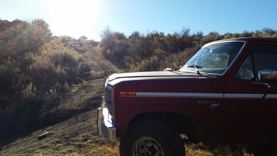 Red 1980s Ford Bronco parked on a dirt road with a hill and sun glare.