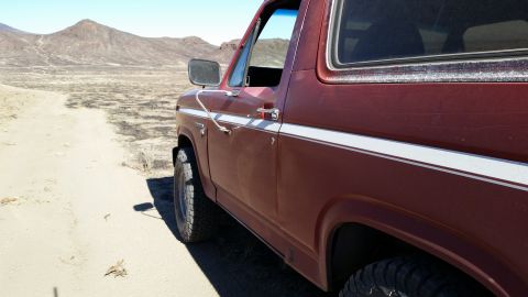 1981 Ford Bronco - A close-up, side-angle view of a maroon Ford Bronco parked on a sandy, desert-like terrain with mountains in the background. The vehicle fe…