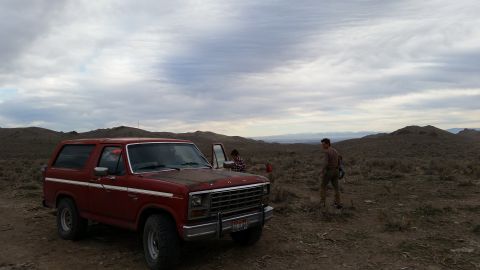 1981 Ford Bronco - A red 1980s Ford Bronco is parked on a dirt road in a dry, hilly landscape under a cloudy sky. Two people are visible near the vehicle, one…