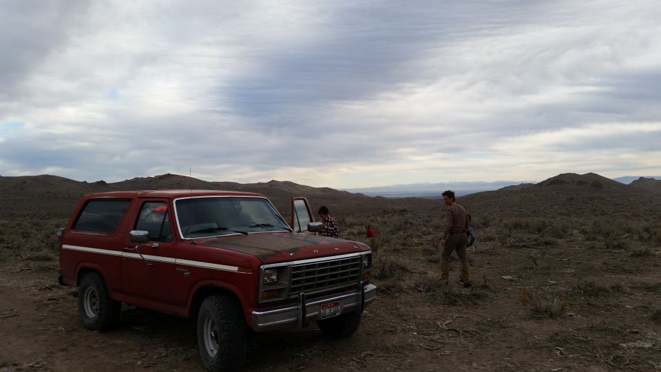 Red 1980s Ford Bronco parked in a dry, hilly landscape with two people nearby.
