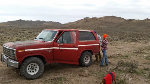 1981 Ford Bronco - A red 1980s Ford Bronco is parked in a dry, arid landscape with rolling hills in the background. Three people and a dog wearing a red vest…