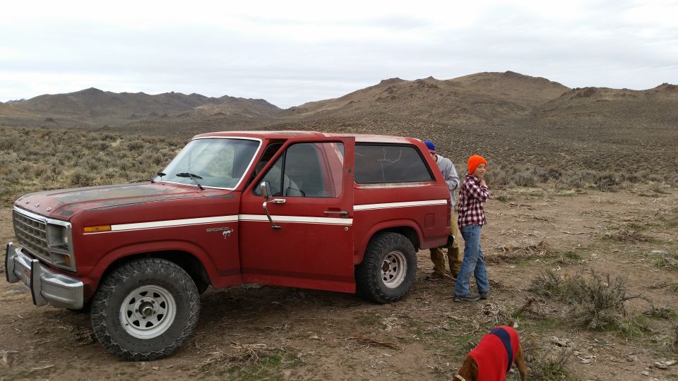 Red 1980s Ford Bronco parked in a dry, arid landscape with people and a dog nearby.