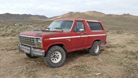 1981 Ford Bronco - A red 1980s Ford Bronco is parked on a dirt road in a dry, hilly landscape under an overcast sky. The vehicle shows some wear, with faded p…