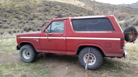 1981 Ford Bronco - A red 1980s Ford Bronco is parked on a dirt road with a dry, hilly landscape in the background. A rifle rests on the ground in front of the…