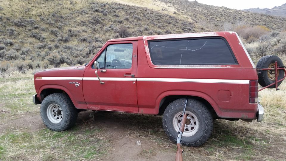 Red 1980s Ford Bronco parked outdoors with a rifle in the foreground.