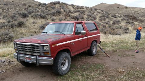 1981 Ford Bronco - A red 1980s Ford Bronco is parked on a dirt road in a dry, hilly landscape. A young boy wearing an orange hat stands to the right of the ve…