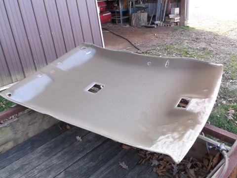 1999 Mercury Grand Marquis - A tan headliner is laid out on a wooden surface, likely a trailer, with a corrugated metal wall in the background. The headliner has cutout…