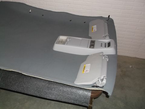 1999 Mercury Grand Marquis - A grey headliner is laid out on a table, with a light grey overhead console and two sun visors attached. The overhead console has buttons a…