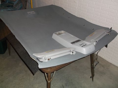 1999 Mercury Grand Marquis - A gray headliner is laid out on a table with two sun visors and a dome light assembly. The headliner has several small cutouts and two squa…