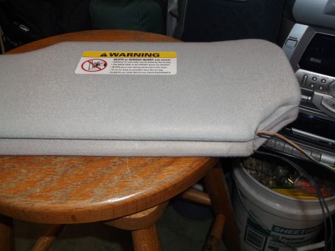 1999 Mercury Grand Marquis - A light gray sun visor with a yellow and red warning sticker is resting on a wooden table. The sticker warns about death or serious injury…