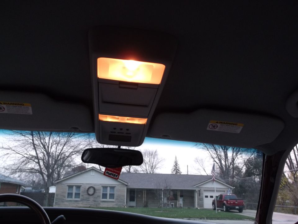Car's overhead console lights are on. Sun visors are down. A house is visible through the windshield