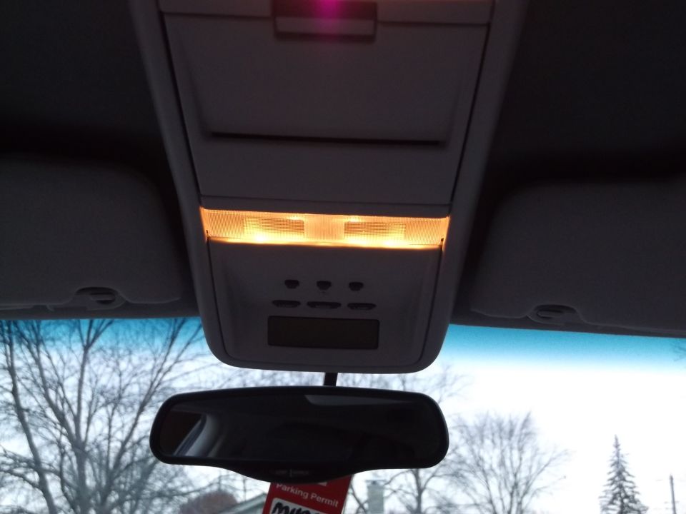 Car's overhead console with lights on, rearview mirror, and a parking permit hanging from it. Trees and sky visible through the windshield