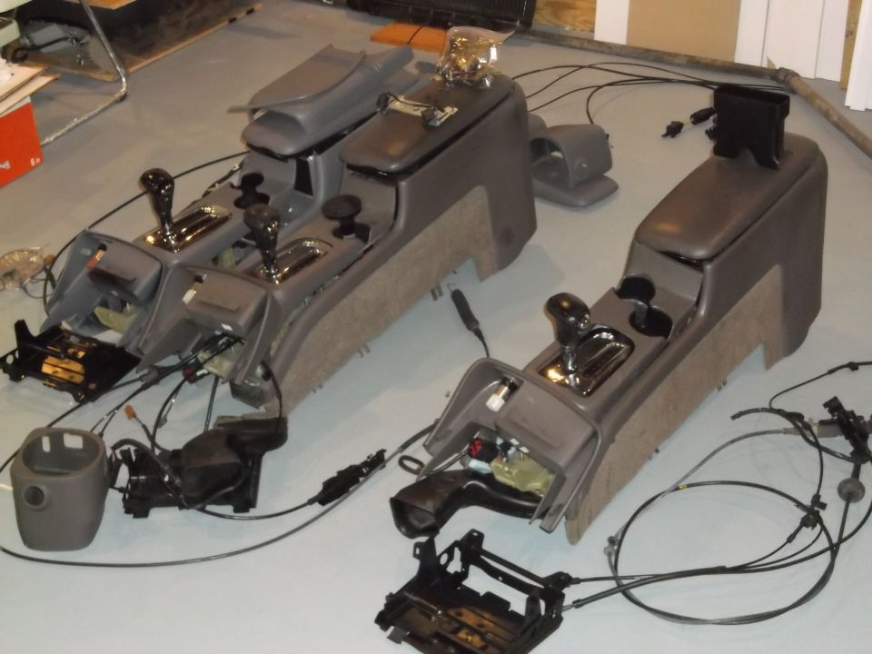 Two disassembled car center consoles with gear shifters and wiring harnesses on a floor