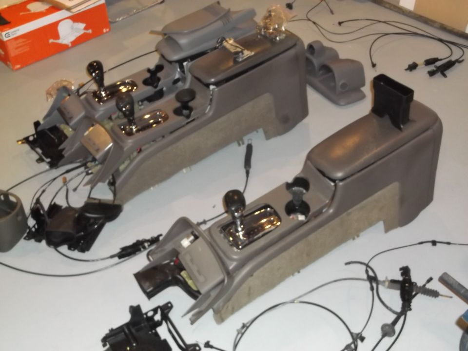 Two disassembled car center consoles with gear shifters and wiring harnesses on a light-colored floor