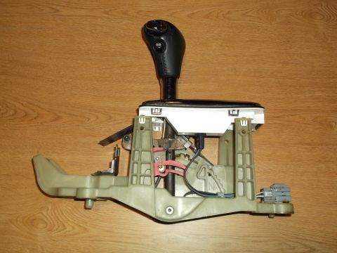 1999 Mercury Grand Marquis - A disassembled automatic transmission shifter assembly is shown from a side angle, resting on a wooden surface. The assembly includes a lea…