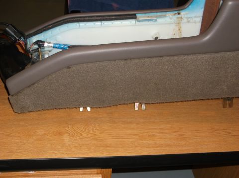 1999 Mercury Grand Marquis - A close-up view of the side of a car's center console, upholstered in brown carpet and grey plastic trim, rests on a wooden surface.