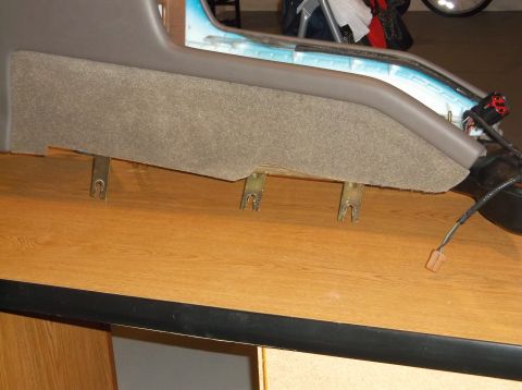 1999 Mercury Grand Marquis - A close-up view of the underside of a car's center console trim panel, showing metal clips and wiring harnesses attached to the gray fabric…